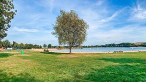 En park med ett stort träd, gräsområden, en sandstrand och en sjö.