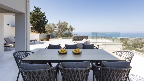 A modern outdoor dining area with a table and chairs, a view of the sea, and a clear sky. - Santa Irini Villas - With 3 Heated Pools (Agia Eirini)