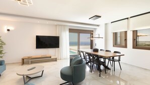A modern living room with a flat-screen TV, a dining area with a table and chairs, a sofa, and a large window offering a view. - Santa Irini Villas - With 3 Heated Pools (Agia Eirini)