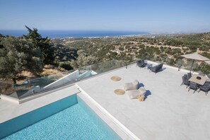 A rooftop pool with a glass balustrade, outdoor seating, and a view of the sea and landscape.