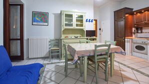 Een eetruimte met een tafel en stoelen, een radiator, een kast met glazen deuren, en een keuken met kasten en apparatuur.