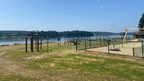 Un parque junto al lago con un área cercada, un conjunto de columpios de madera y un pabellón.