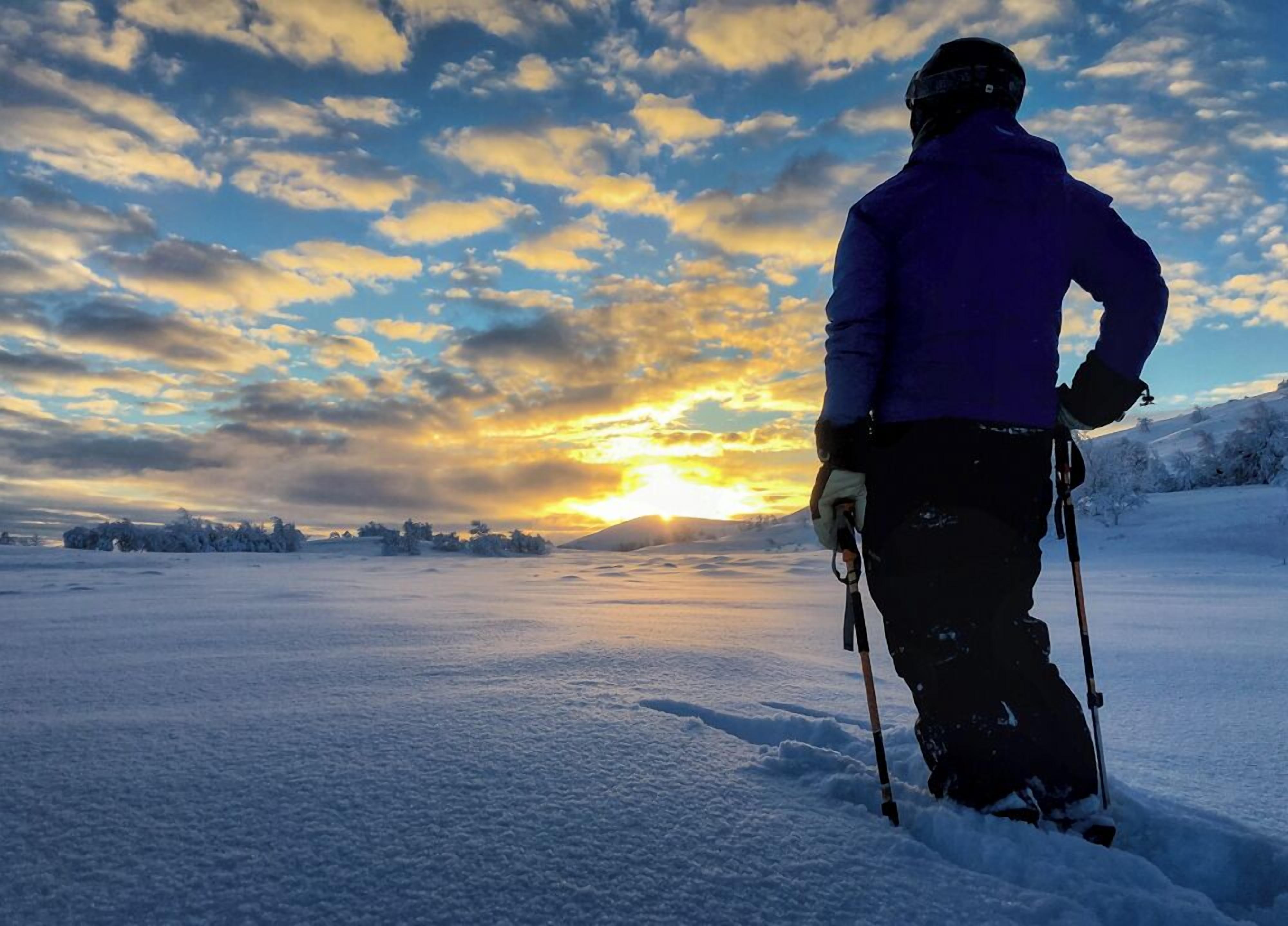 Une personne fait du ski dans un paysage enneigé, avec un coucher de soleil en arrière-plan.