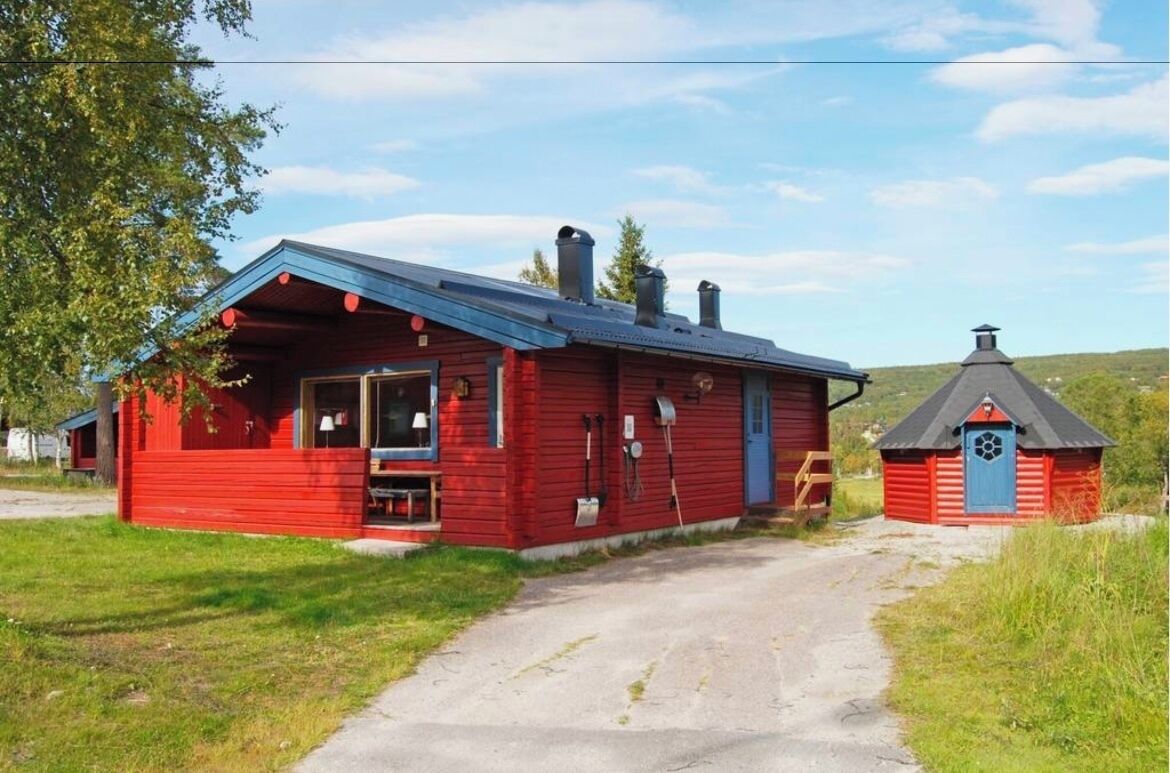 En rød hytte med blå tag, et lille tilbyggeskur og en jordvej, der fører til hytten.