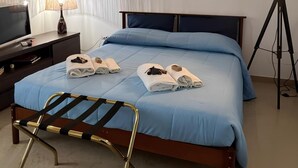 Un lit bien fait, avec une literie bleue, une lampe de chevet, un téléviseur posé sur un meuble en bois et une lampe avec un abat-jour blanc.