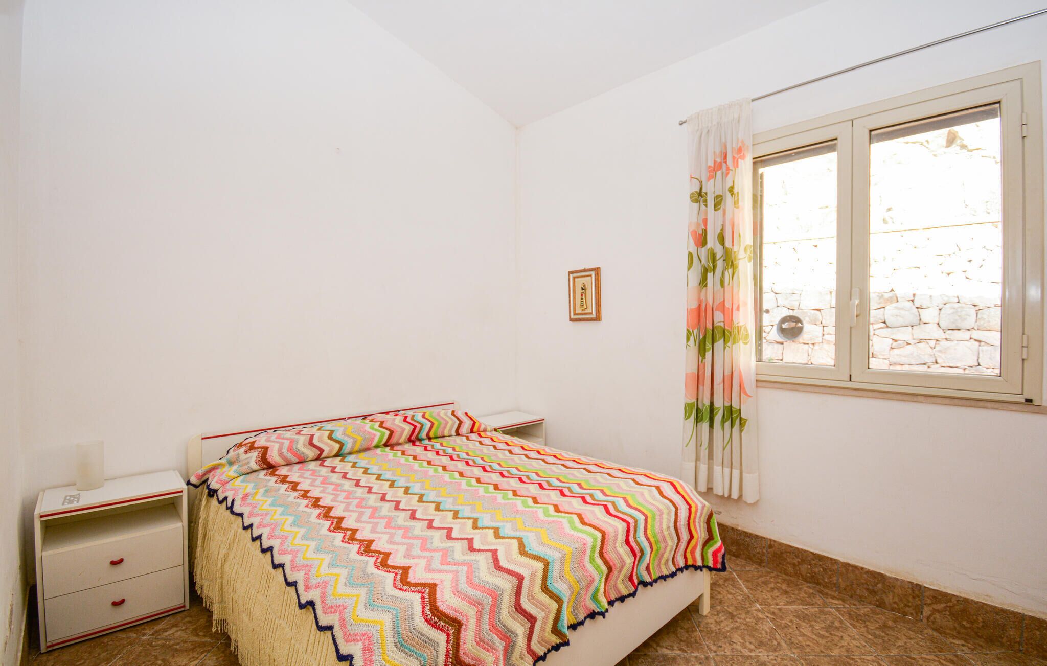 Une chambre à coucher avec un lit, une table de chevet, une fenêtre avec des rideaux et un tableau encadré au mur.