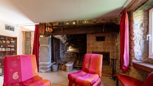 Una habitación acogedora con chimenea de piedra, dos sillones con tapicería estampada, un estante de madera con adornos y una ventana con cortinas.