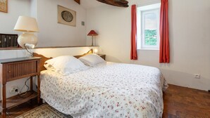Un dormitorio con cabecera de madera, cama con ropa de cama floral, mesita de noche con lámpara, ventana con cortinas rojas y vista a follaje exterior.