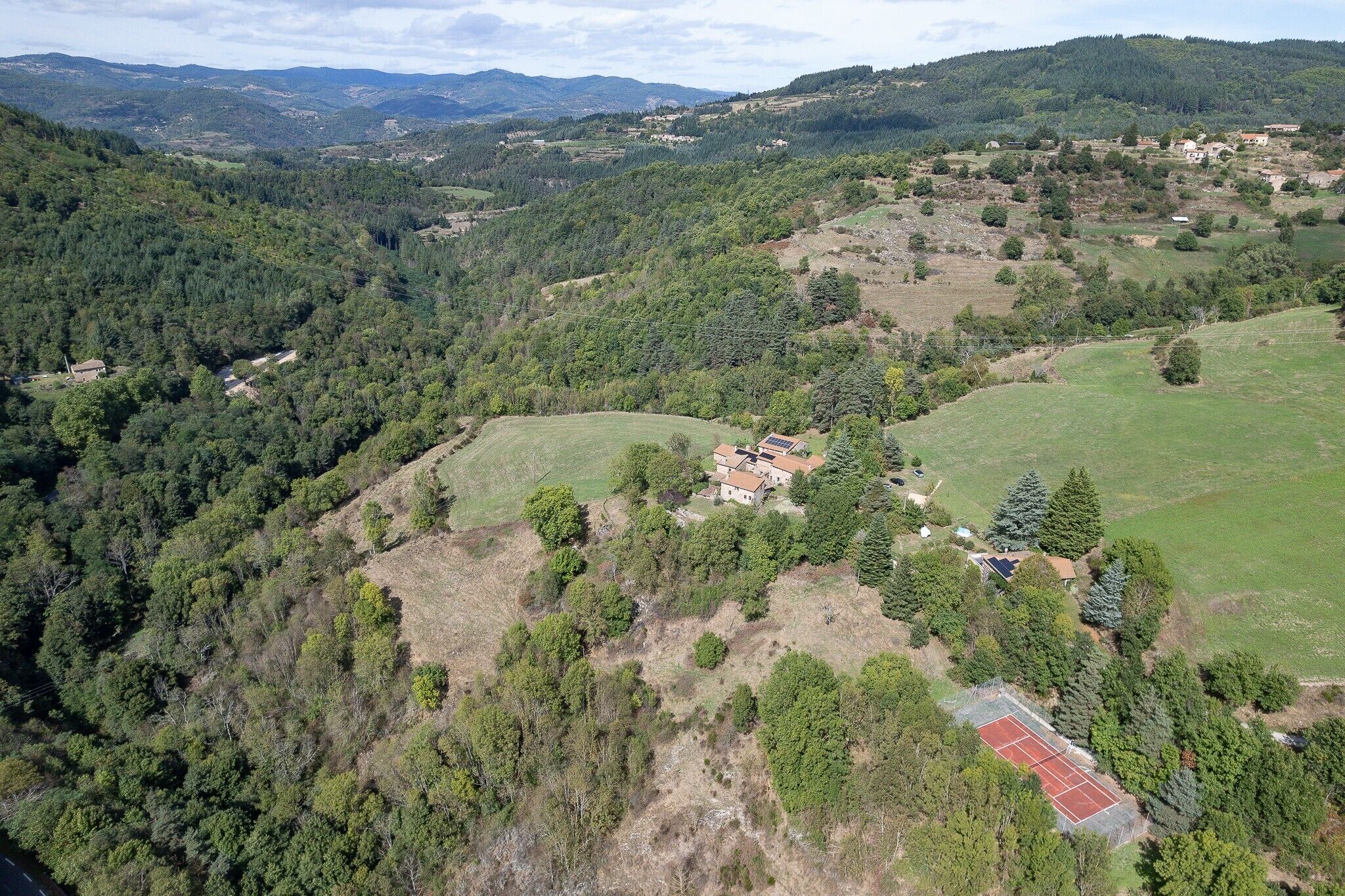 An aerial view of a rural area with a tennis court, scattered houses, and dense forested hills.