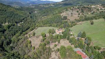 An aerial view of a rural area with a tennis court, scattered houses, and dense forested hills.