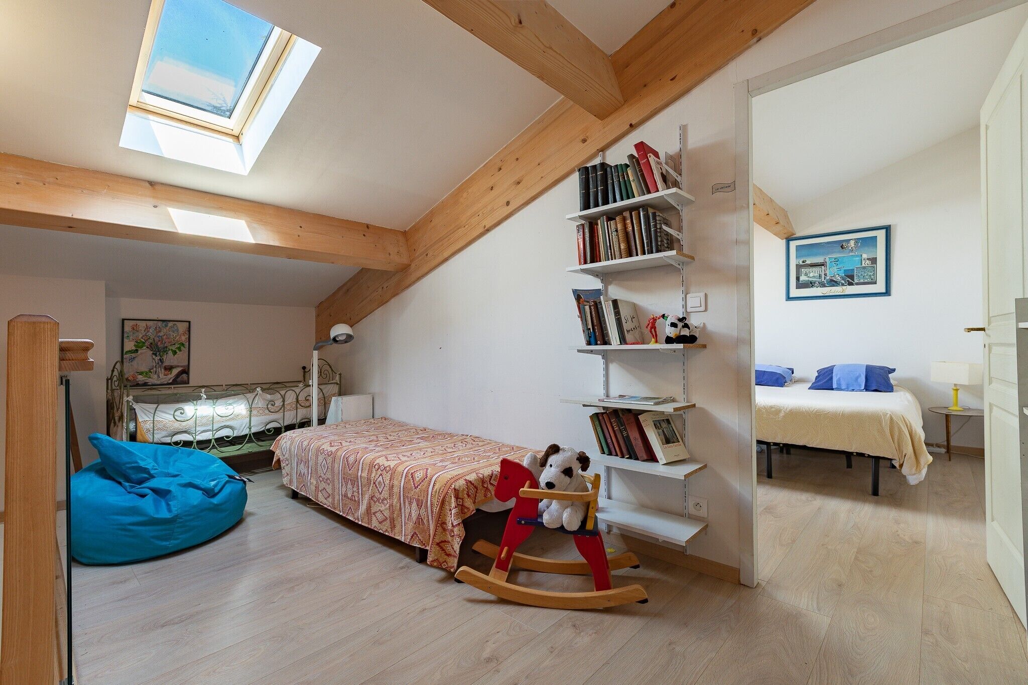 A room with a sloped ceiling, a skylight, a bed with a patterned bedspread, a rocking chair with a stuffed animal, a bookshelf, and a painting on the wall.