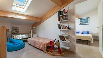 A room with a sloped ceiling, a skylight, a bed with a patterned bedspread, a rocking chair with a stuffed animal, a bookshelf, and a painting on the wall.