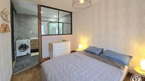 A bedroom with a large bed, a mirror, a washing machine, and a bedside table with a lamp.