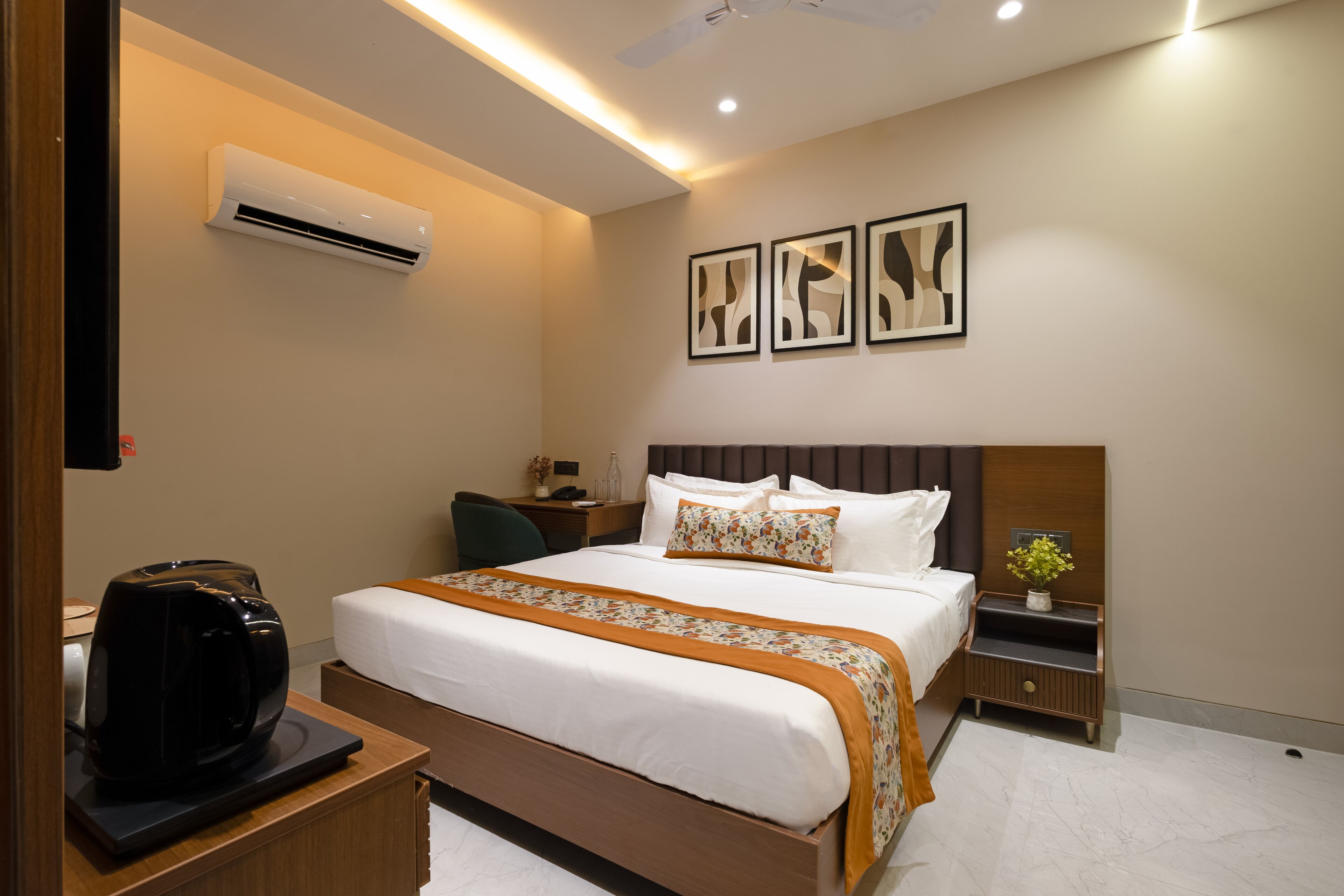 A modern hotel room with a neatly made bed, a bedside table with a coffee maker, a wall-mounted air conditioner, and a small nightstand with a vase of flowers.