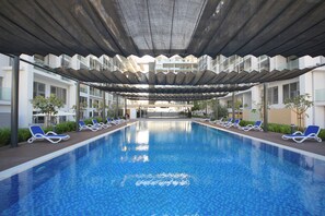 A swimming pool with blue tiled floor, surrounded by lounge chairs and a covered area with a modern architectural design. - Silk Valley-Cozy Studio near Masdar Park (Abu Dhabi)