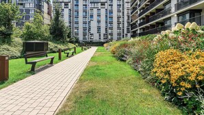 Ein Weg führt zu einem modernen Mehrfamilienhaus mit mehreren Balkonen und einem gepflegten Garten mit gelben Blumen.
