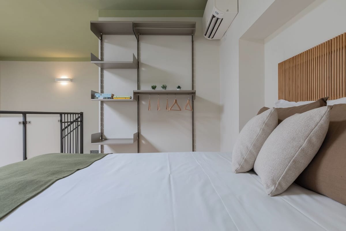 A modern bedroom with a bed, a wooden headboard, a shelf with books and plants, and a staircase.