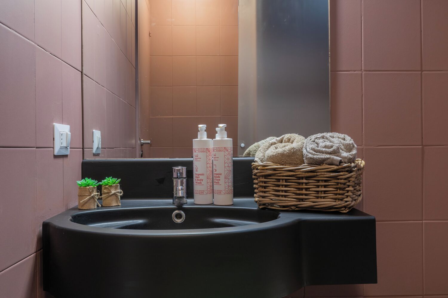 A bathroom with a black sink, two bottles of organic hair and body wash, a small plant, and a wicker basket with towels.