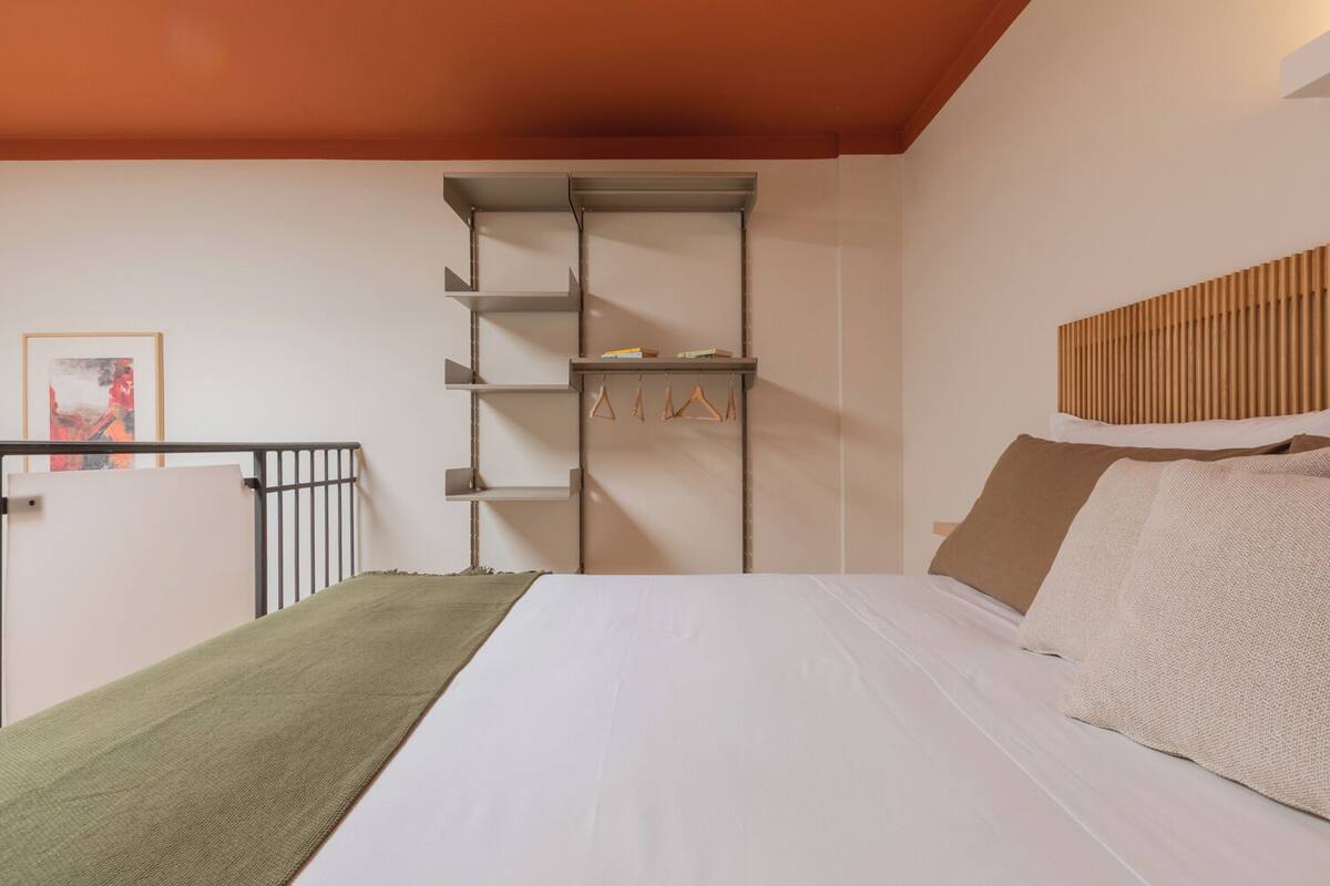 A bedroom with a bed, a metal shelf unit, a wooden headboard, and a painting on the wall.