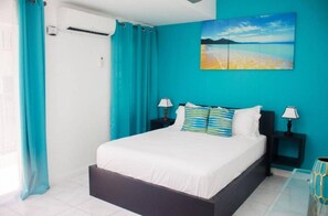 A bedroom with a bed, two bedside tables with lamps, a window with curtains, and a beach scene painting on the wall.
