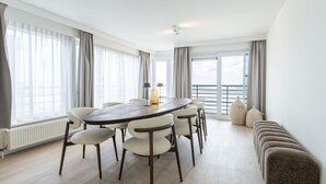 Un comedor moderno con una mesa y sillas de madera, ventanas amplias con cortinas y vistas al mar.