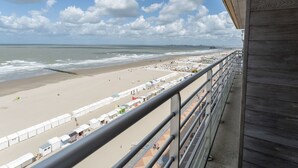 Vista a la playa desde un balcón con barandilla de madera, una playa de arena y un muelle a lo lejos.