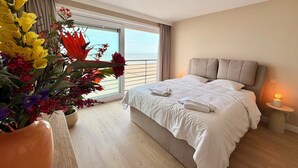 Una habitación de hotel con una cama grande, una mesita de noche con una lámpara, una planta en maceta con flores y vista al océano a través de un ventanal amplio.