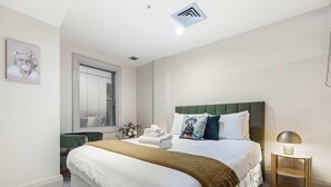 A bedroom with a large bed, a bedside table with a lamp, a window with blinds, and a wall-mounted picture. - Comfort, Harbour Views & Balcony | Zodiak Stays (Auckland)