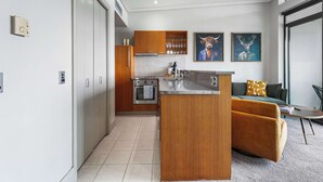A modern kitchen with wooden cabinets, a central island, and a built-in oven. There is a sofa with a patterned cushion and a coffee table in the living area. Two framed artworks are on the wall. - Comfort, Harbour Views & Balcony | Zodiak Stays (Auckland)