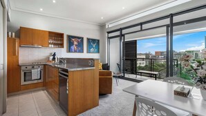 A modern kitchen with wooden cabinets, a built-in oven, and a large island. There is a dining area with a table and chairs, and a balcony with a view of the city. - Comfort, Harbour Views & Balcony | Zodiak Stays (Auckland)