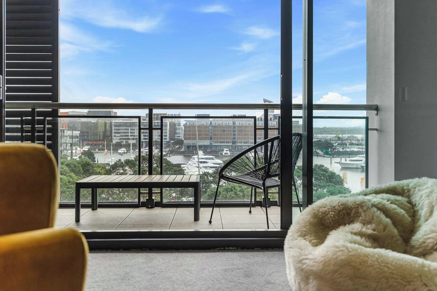 A balcony with a view of a marina and buildings, a wooden table, a black chair, and a fluffy white bean bag.