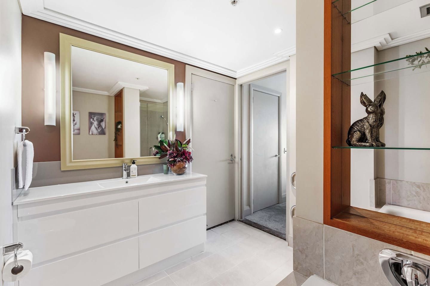 Een moderne badkamer met een grote spiegel, witte kastjes, een decoratieve plant en een glazen plank met een beeldje.