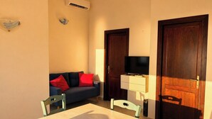 A room with a wooden table, two chairs, a sofa with red cushions, a television on a stand, and two dark wooden doors.