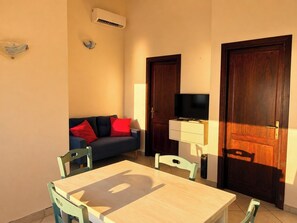 Une pièce avec une table en bois, deux chaises, un canapé avec des coussins rouges, un téléviseur posé sur un meuble et deux portes en bois foncé.
