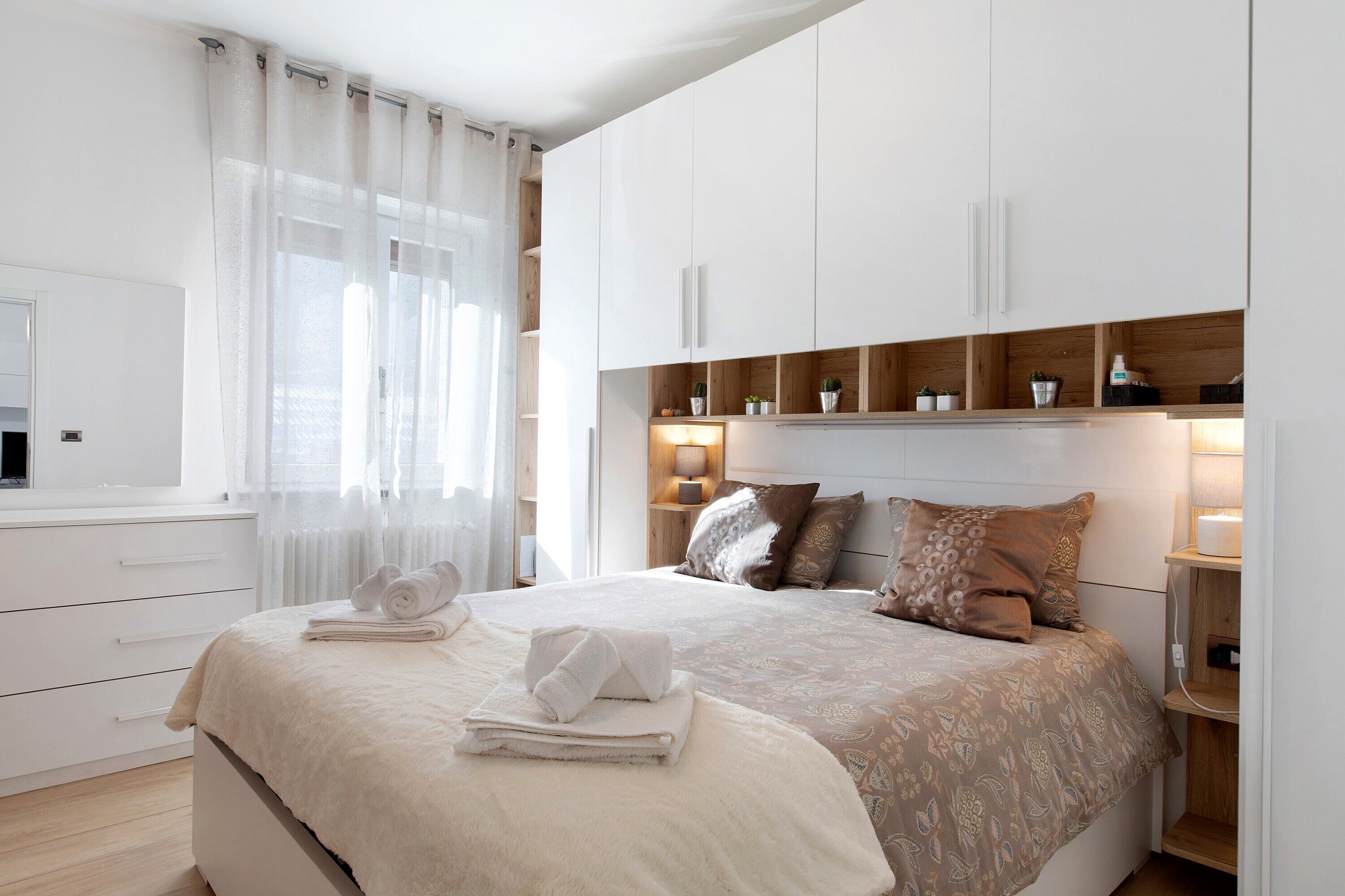 A bedroom with a bed, a bedside table, a nightstand, a window with sheer curtains, and a wall-mounted shelf with decorative items.