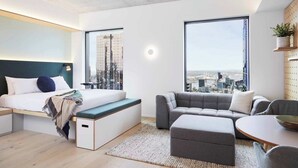 A modern bedroom with a bed, bench, couch, and a city view through large windows.