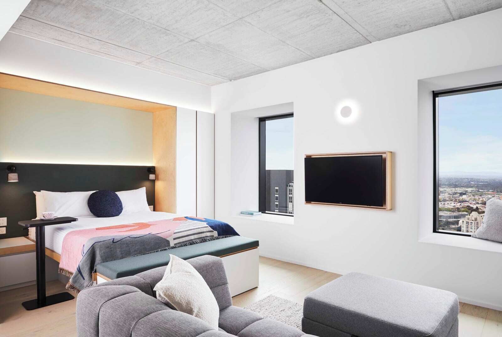 Une chambre d’hôtel moderne dotée d’un grand lit, d’un bureau, d’une chaise, d’un canapé et offrant une vue sur la ville grâce à une grande fenêtre.