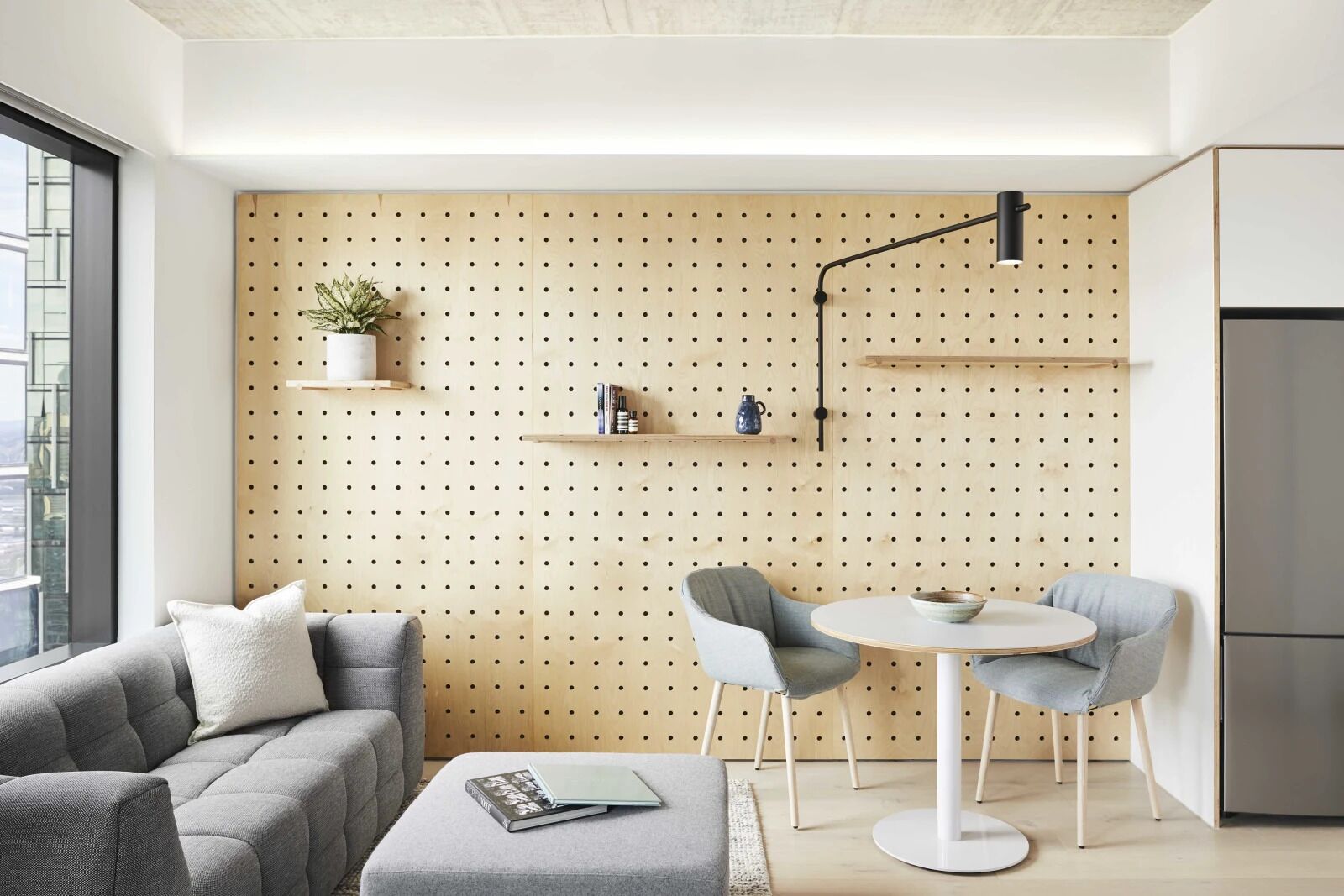 Una sala de estar moderna con un sofá, una mesa redonda y sillas. Se observa una pared de madera con estantes y una planta colgante.
