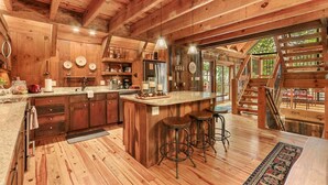 A rustic wooden cabin interior with a kitchen, bar area, and a staircase leading to a deck.