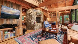 A cozy cabin interior with a stone fireplace, wooden furniture, a large flat-screen TV, and a stone wall.