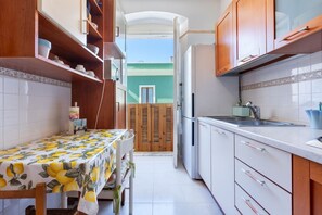 A narrow kitchen with white cabinets, a wooden table with a lemon tablecloth, and a view through an open door to a balcony.