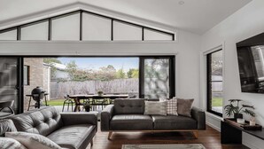 A modern living room with a leather sofa, a dining table with chairs, and a large window overlooking a backyard. - Luxury On Lakeside (Mount Beauty)