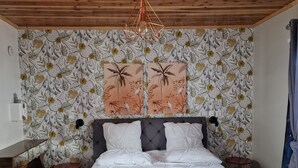 Un dormitorio con techo de madera, cama con edredón blanco, papel tapiz floral y lámpara colgante.
