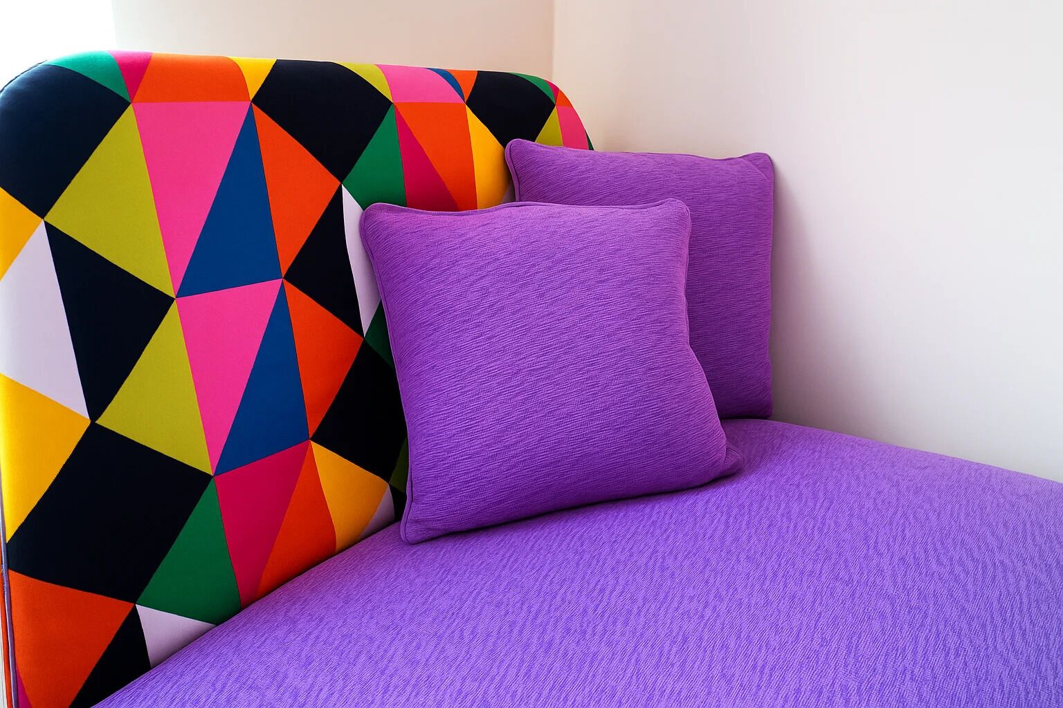 Een hoek van een kamer met een kleurrijk hoofdeinde met een geometrisch patroon en twee paarse kussens.