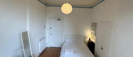 Et soveværelse med trægulv, en seng, et sengebord med lampe, en kommode, et spejl lænet op ad væggen og en dør, der fører til et andet rum.
