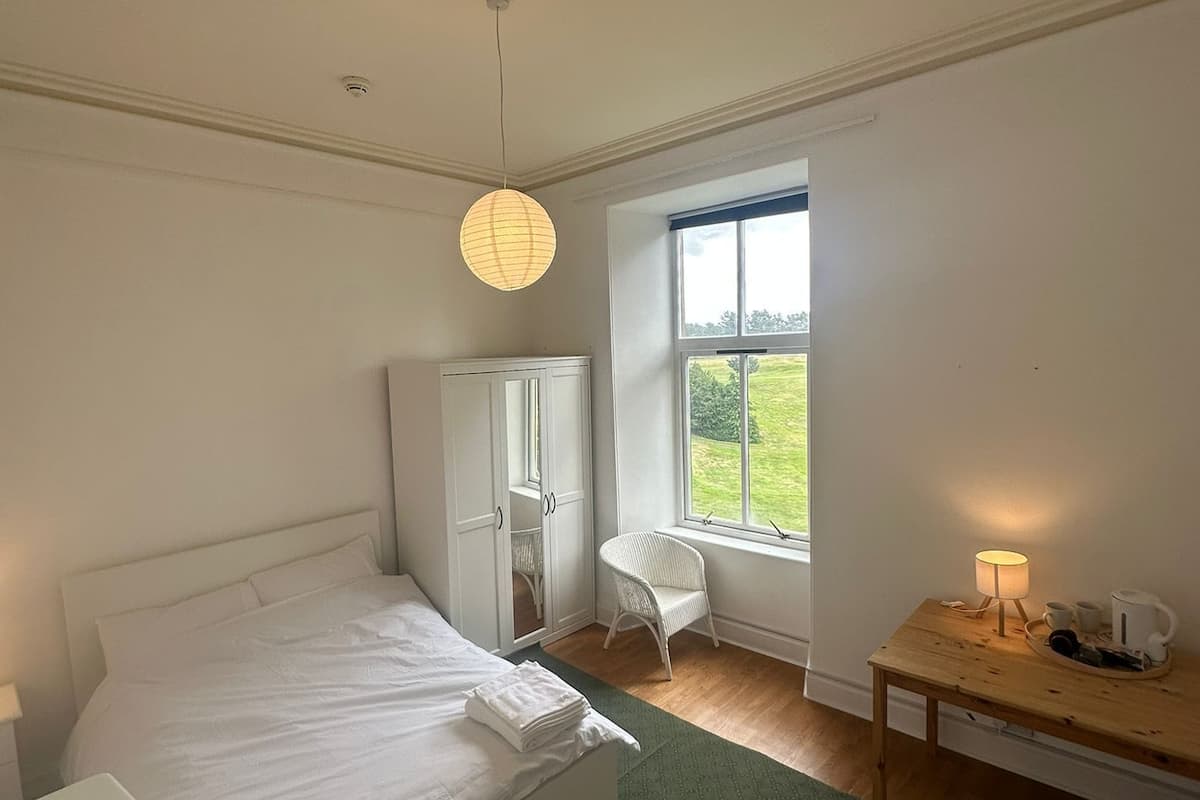 A bedroom with a bed, a chair, a table with a lamp, and a window with a view of greenery.