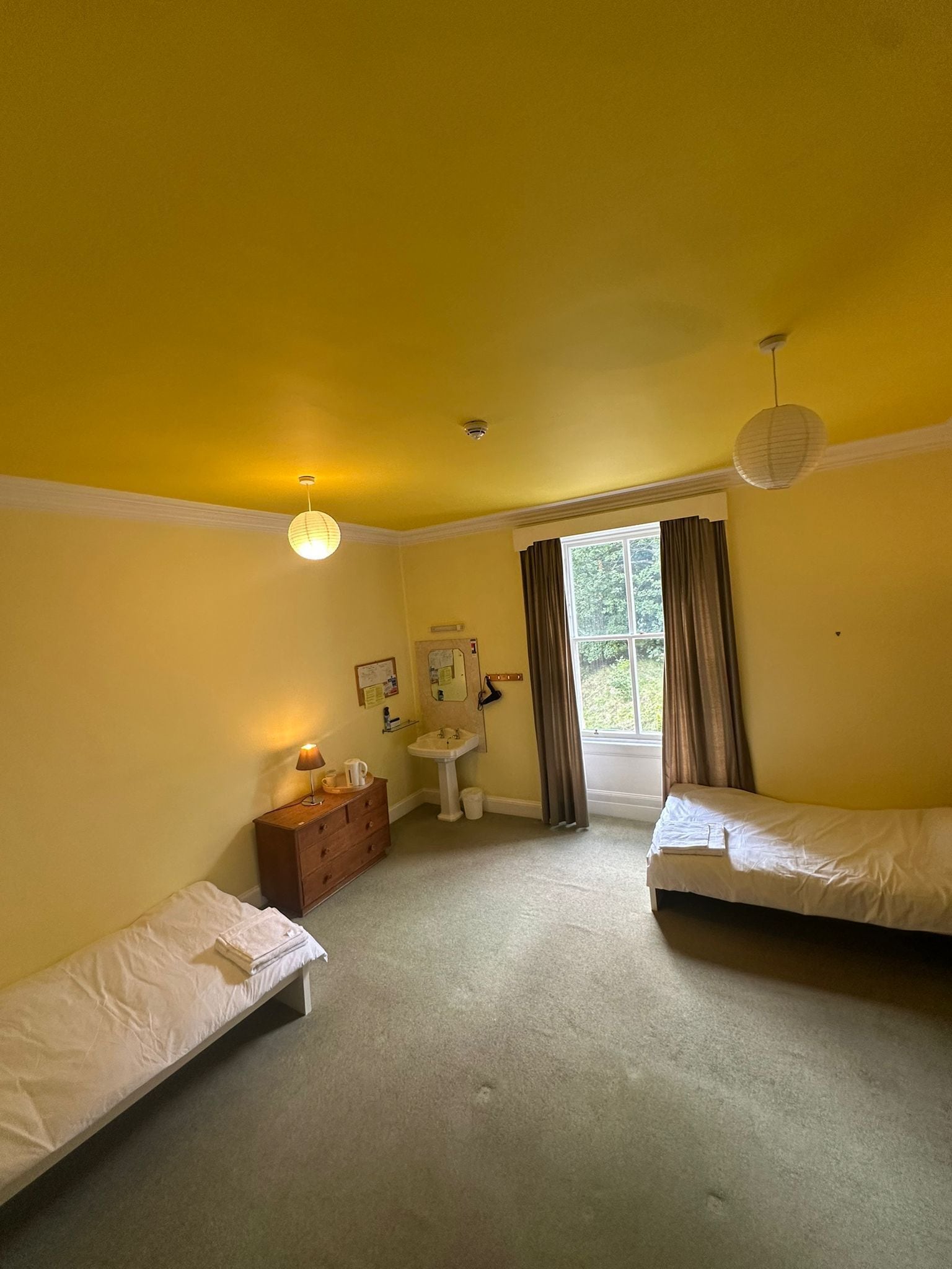 A room with two single beds, a wooden dresser, a sink, and a window with curtains.