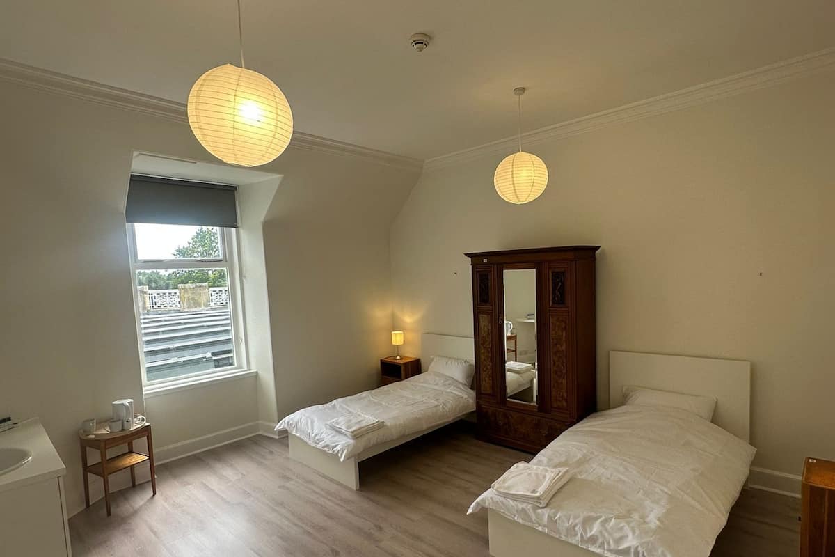 A bedroom with two single beds, a wooden wardrobe, a small bedside table with a lamp, and a window with blinds.