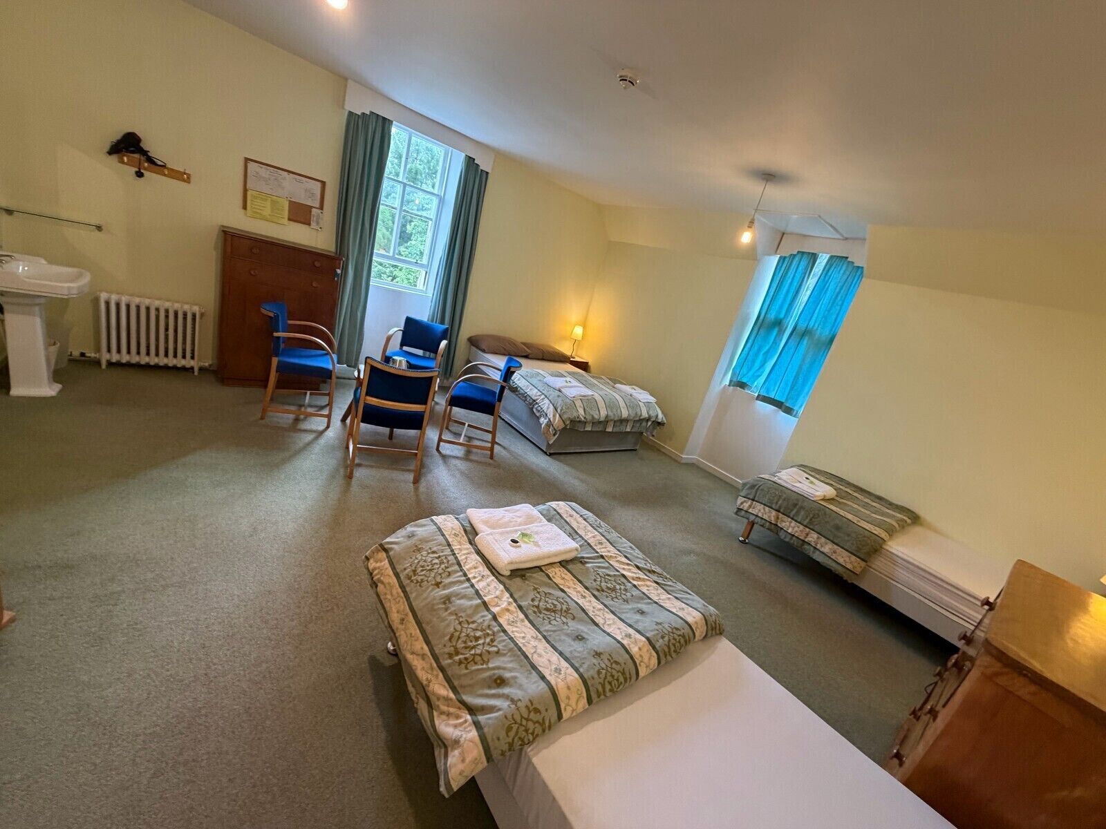 A hotel room with a bed, two chairs, a desk, a radiator, a window with curtains, and a ceiling fan.