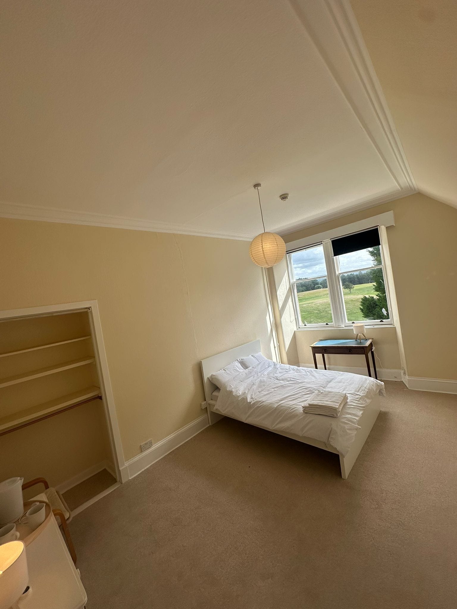 A bedroom with a single bed, a window with a view of a green landscape, a small table with a lamp, and a wardrobe.